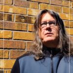 image of Isaac Gruzinsky. Male with shoulder length hair wearing zipped up sports hoodie. background is a brick wall