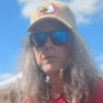 Isaac is outdoors at a concert. image is a head shot. Isaac is wearing a bill cap with eagle patch on it. he is also wearing sunglassses.